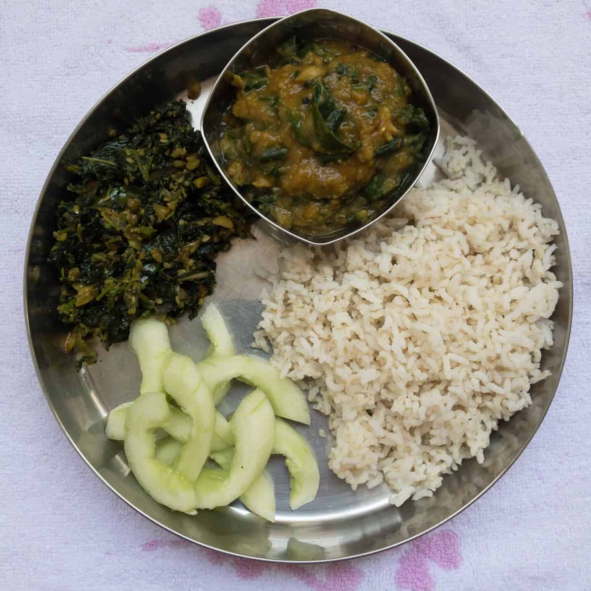 Healthy lunch plate with rice, kale dal, leafy stir-fry, and cucumber slices.