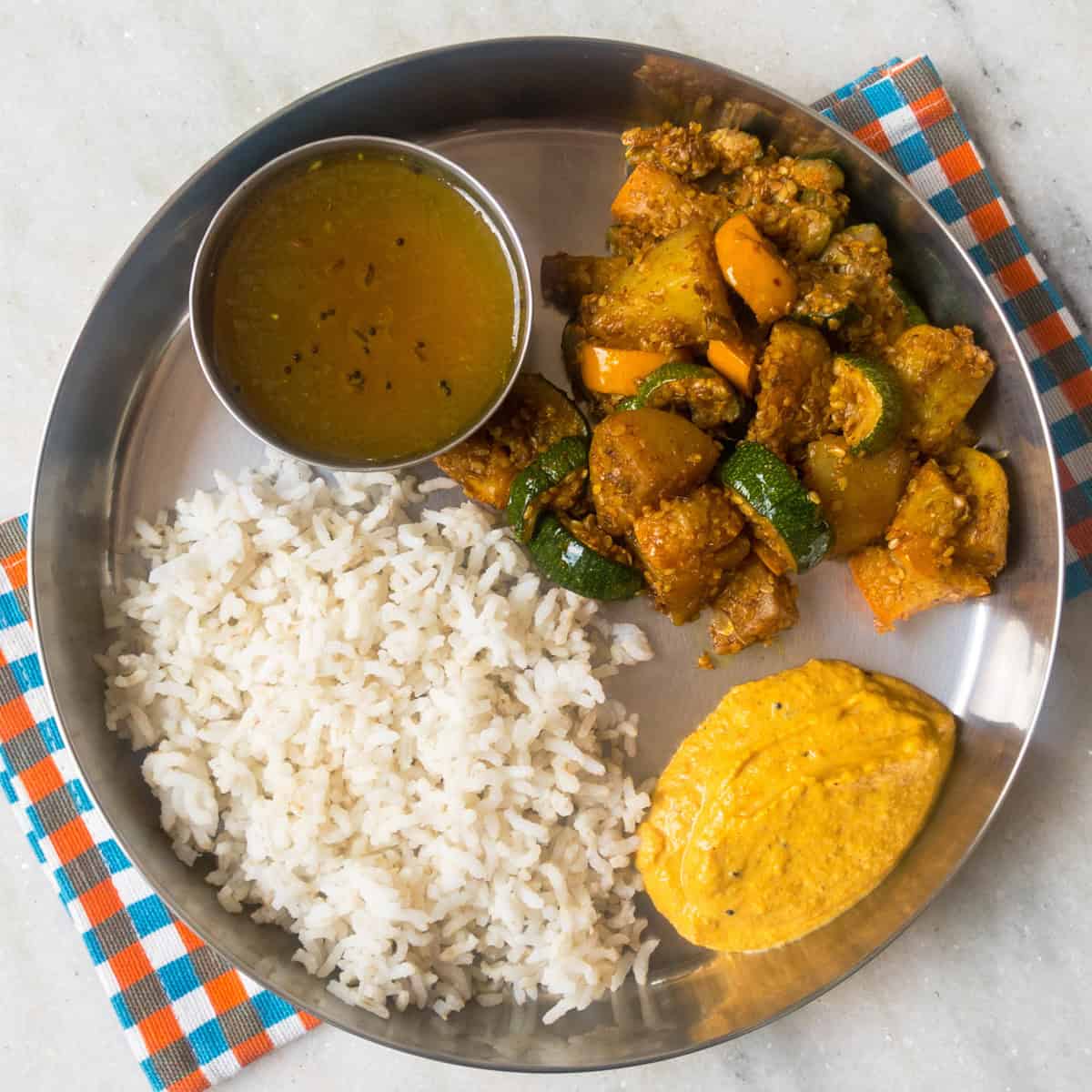 Indian lunch plate with rice, rasam, zucchini and potato sabzi, and tomato chutney.