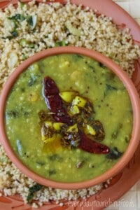 A bowl of gongura pappu topped with tempering of dried red chilies and spices, served on a plate with cooked millet.