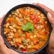 A bowl of colorful vegetable and black eyed peas soup garnished with a cilantro leaf, held by two hands