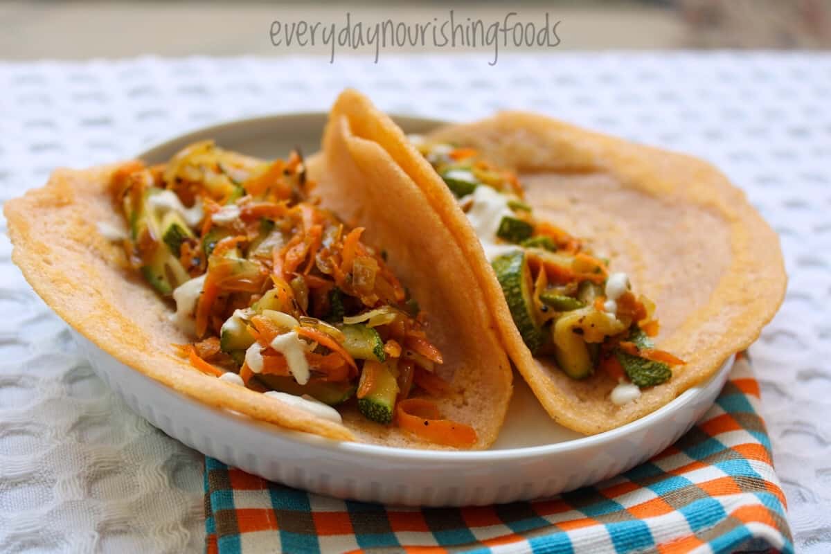 Two gluten-free lentil wraps filled with a colorful mix of sautรฉed carrots, zucchini, and broccoli, garnished with a creamy sauce, served on a white plate with a checkered napkin background.