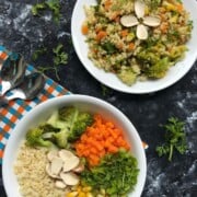 A colorful millet salad served with steamed broccoli, carrots, and corn, topped with fresh herbs, presented in a white bowl on a wooden table.