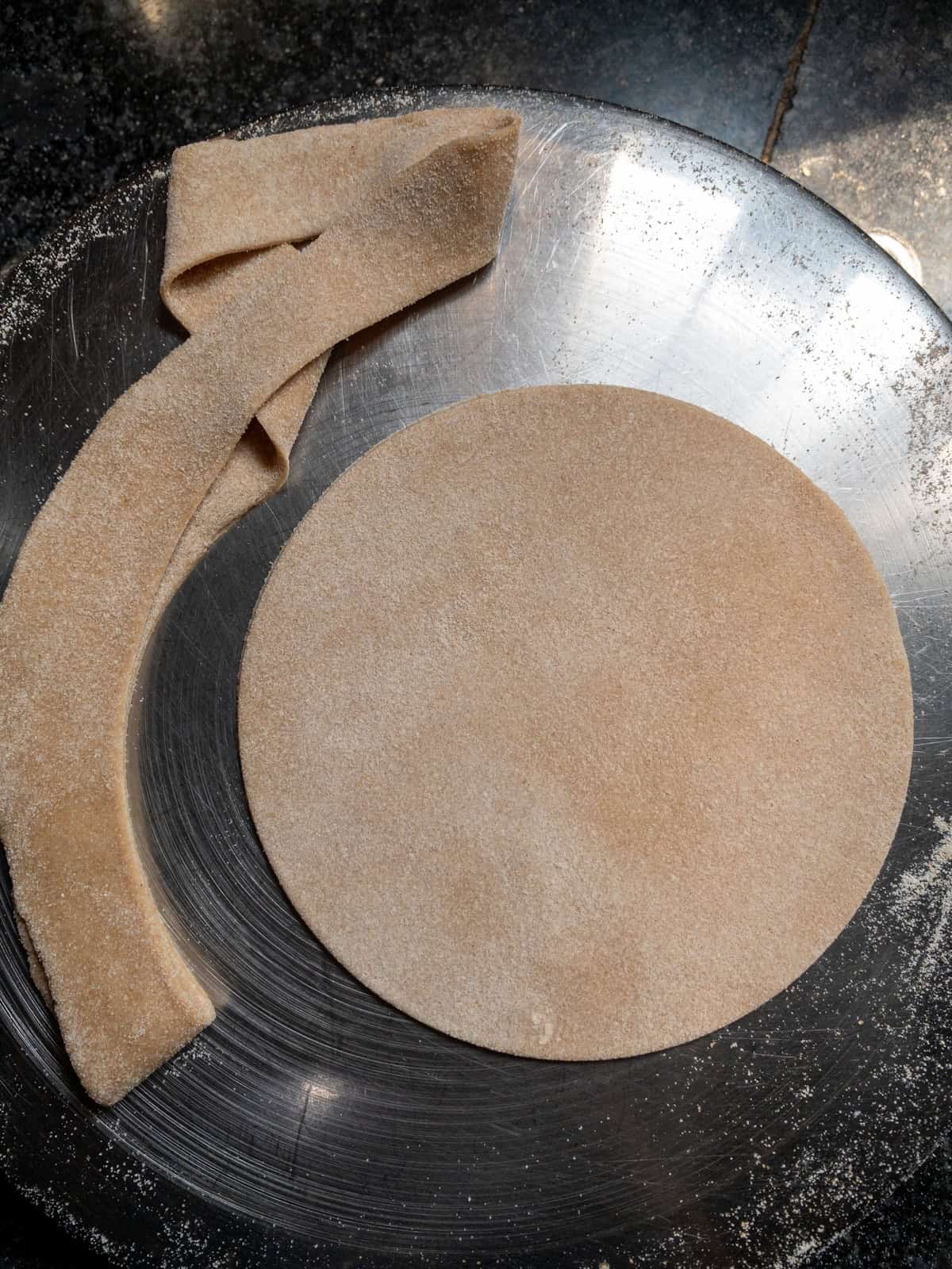 A perfectly round chapati dough disc on a stainless steel plate, with excess dough strips cut off to achieve the circular shape.