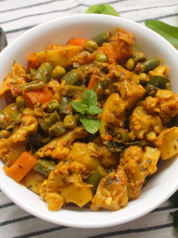 A bowl of Indian vegetable curry garnished with fresh mint, served with a side of curry leaves on a striped napkin.