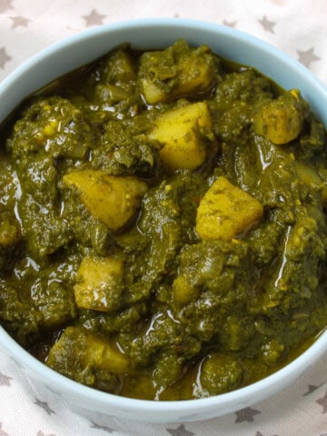 A bowl of Saag Aloo – Indian spinach and potato curry – served in a light blue bowl, showing chunky potatoes coated in a vibrant green spinach gravy.