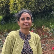 Sravanthi Chanda standing in a rose garden, wearing a light green embroidered sweater and patterned black kurta, smiling softly with trees and flowers in the background.