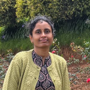 Sravanthi Chanda standing in a rose garden, wearing a light green embroidered sweater and patterned black kurta, smiling softly with trees and flowers in the background.