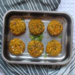 Six golden-brown millet black eyed peas patties arranged in a steel tray with fresh cilantro garnish.