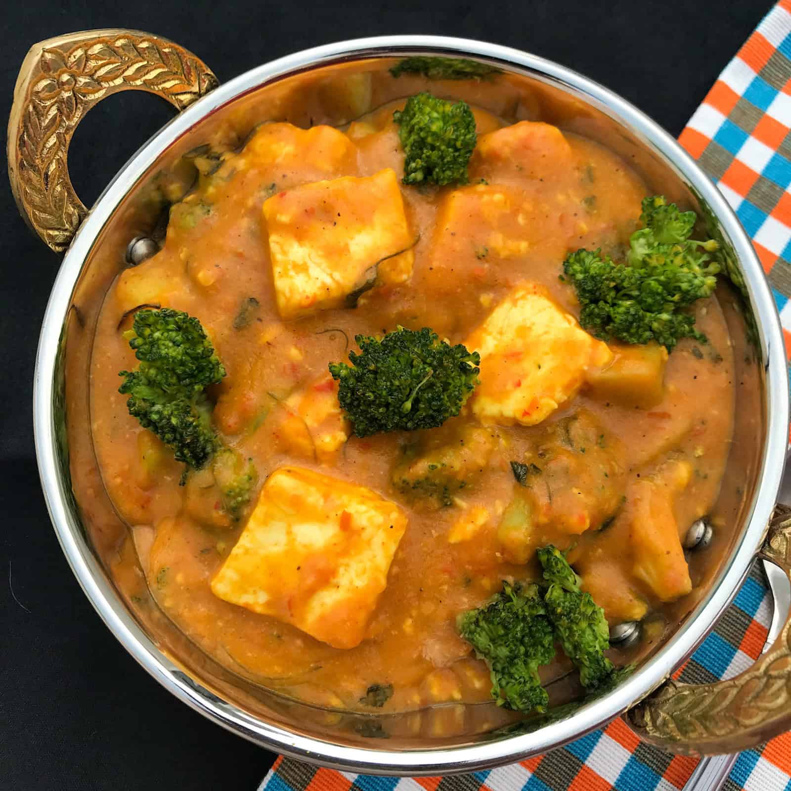 Close-up of broccoli paneer curry in a brass-handled serving bowl, showing golden paneer cubes and broccoli florets in a spiced tomato gravy.