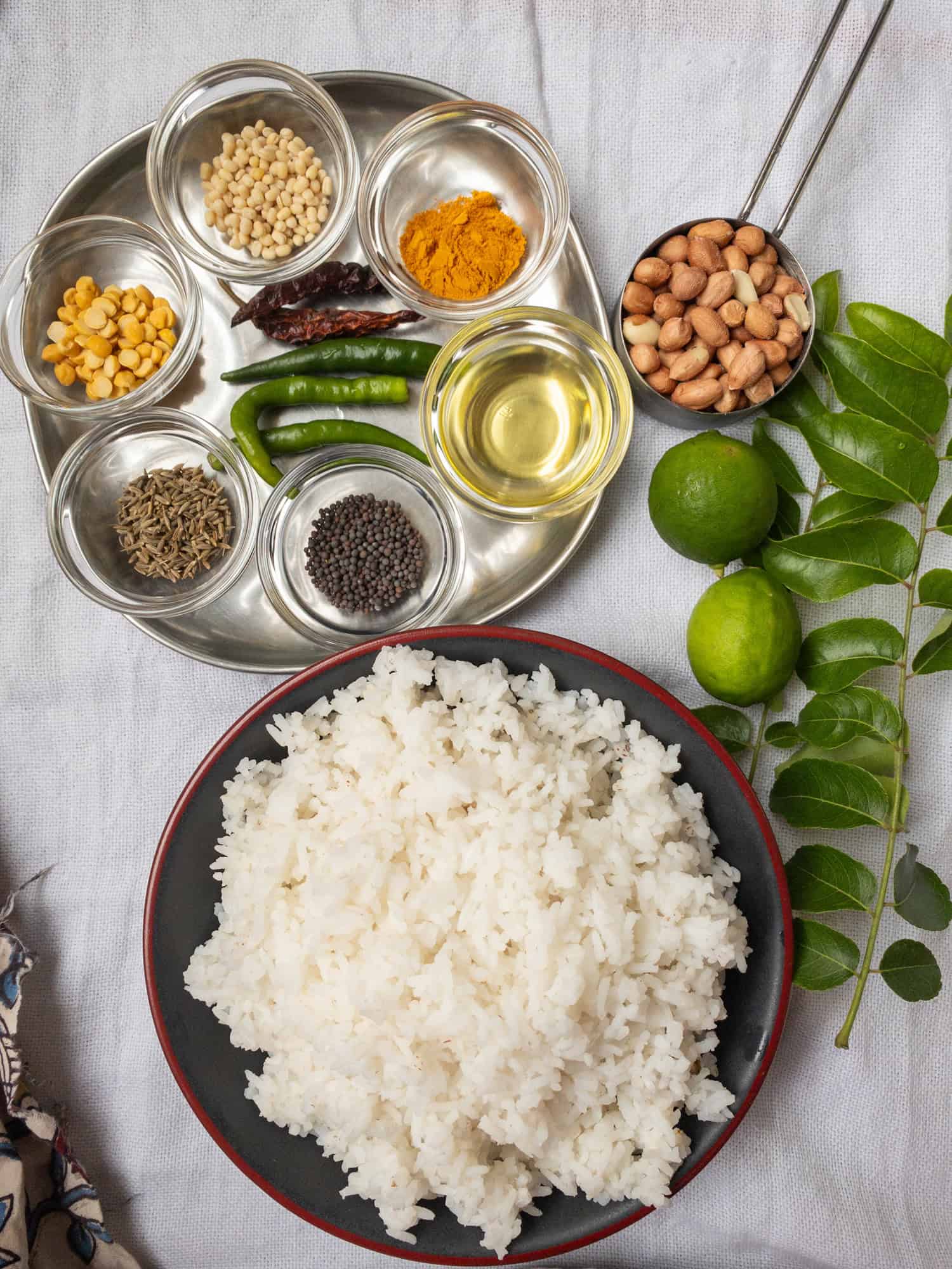 A bowl of cooked basmati rice with a plate of ingredients including peanuts, curry leaves, lemon, chilies, mustard seeds, cumin seeds, chana dal, urad dal, turmeric powder, oil, and dried red chilies.