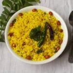 Indian lemon rice in a white bowl topped with fried peanuts, curry leaves, and chilies, placed on a light background with curry leaves on the side.