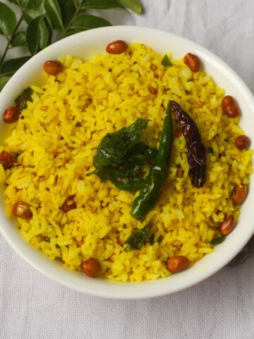 Indian lemon rice in a white bowl topped with fried peanuts, curry leaves, and chilies, placed on a light background with curry leaves on the side.