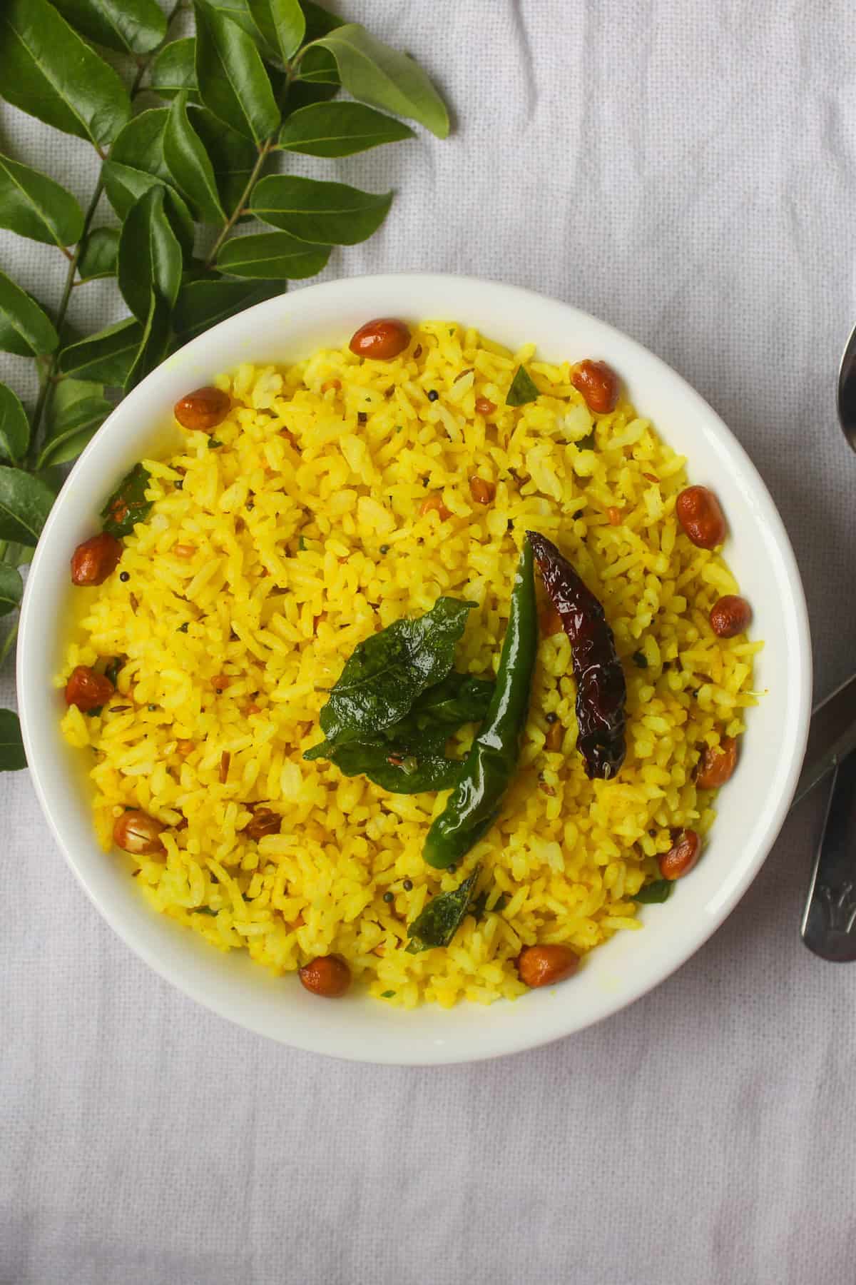 Overhead view of Indian lemon rice with bright yellow grains, roasted peanuts, curry leaves, and chilies, served in a white bowl on a grey cloth.