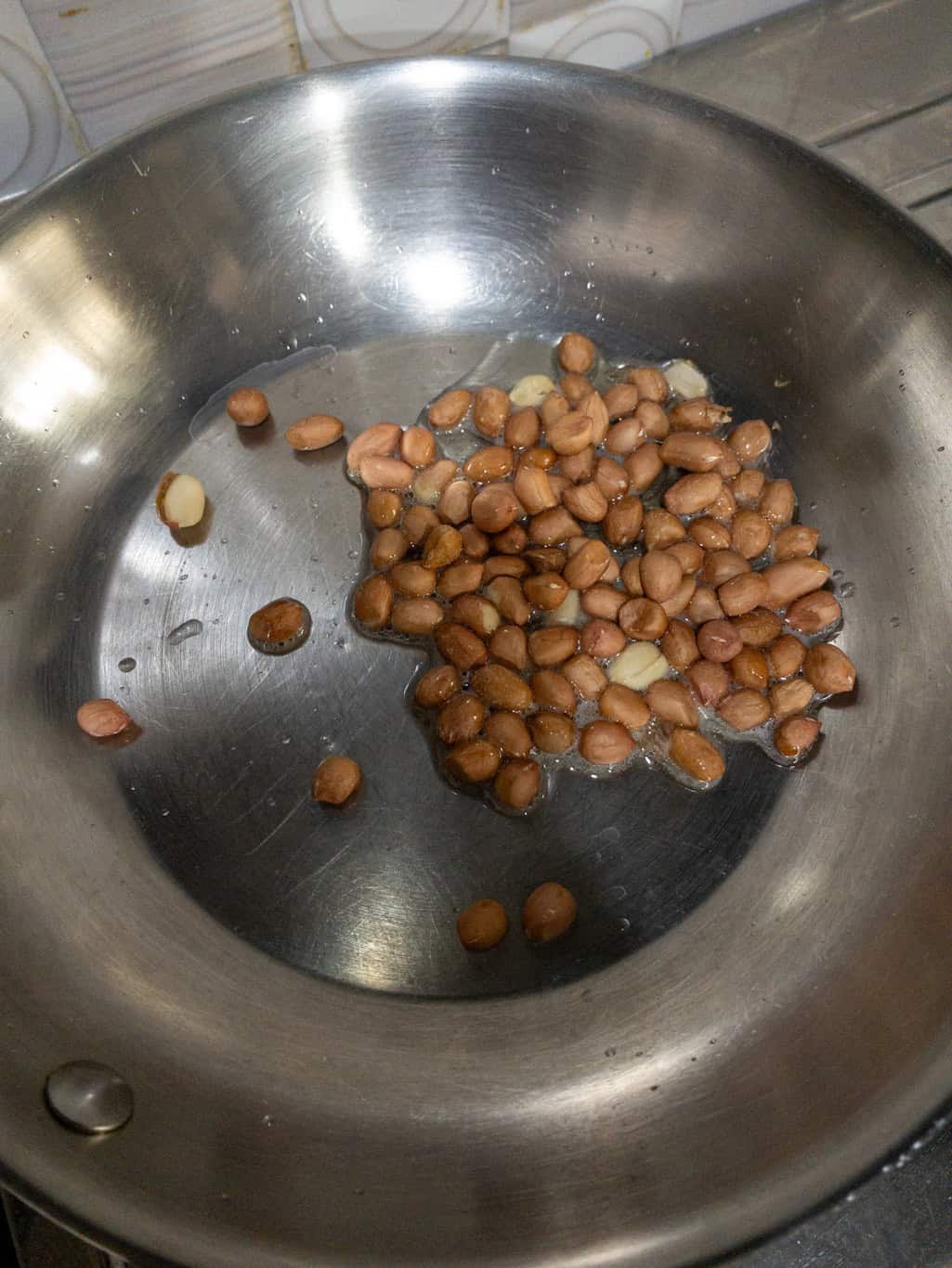 Raw peanuts roasting in hot oil inside a stainless steel pan until golden and crunchy.