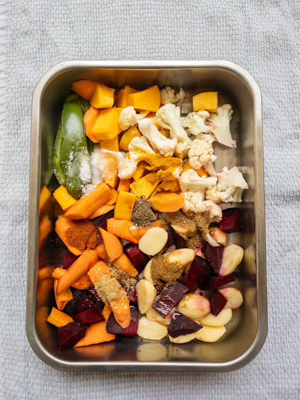 Chopped vegetables like beets, carrots, cauliflower, and peppers with spices and oil in a steel roasting pan.