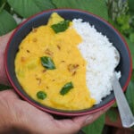 A bowl of creamy yellow moong dal served with steamed rice, garnished with curry leaves and cumin tempering, held in both hands against a green leafy background.