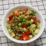 A white bowl filled with Kachumber salad made of cucumber, tomato, onion, and cilantro, garnished with fresh coriander leaves.