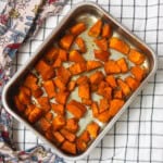 Roasted pumpkin cubes in a steel baking tray, golden brown and spiced, resting on a checkered cloth with a printed kitchen towel.