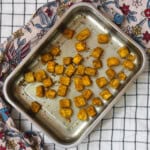 Top view of crispy oven-baked tofu cubes with Indian spices arranged in a steel tray on a patterned cloth.