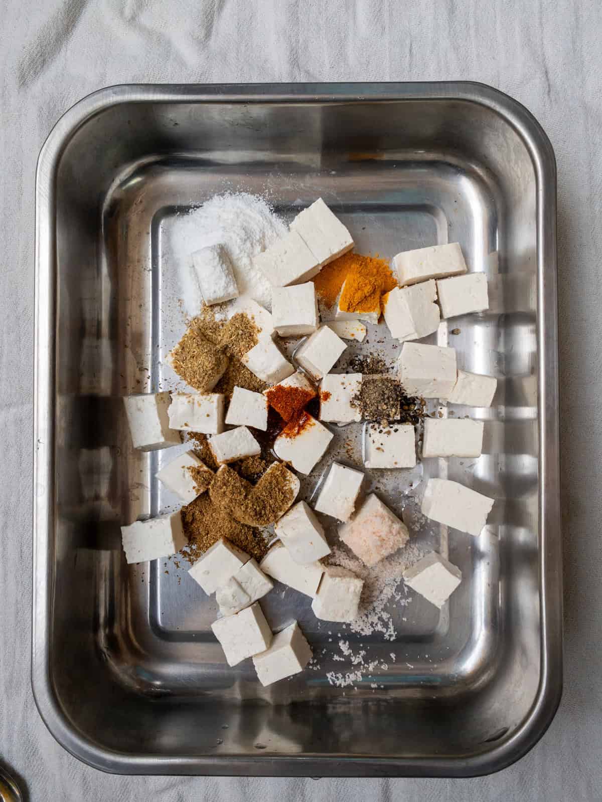 Raw tofu cubes in a tray with turmeric, chili powder, coriander, pepper, salt, and rice flour before mixing.