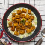 Indian spiced roasted tofu cubes served on creamy Indian yogurt salad with cucumber and tomato in a black bowl, garnished with coriander.