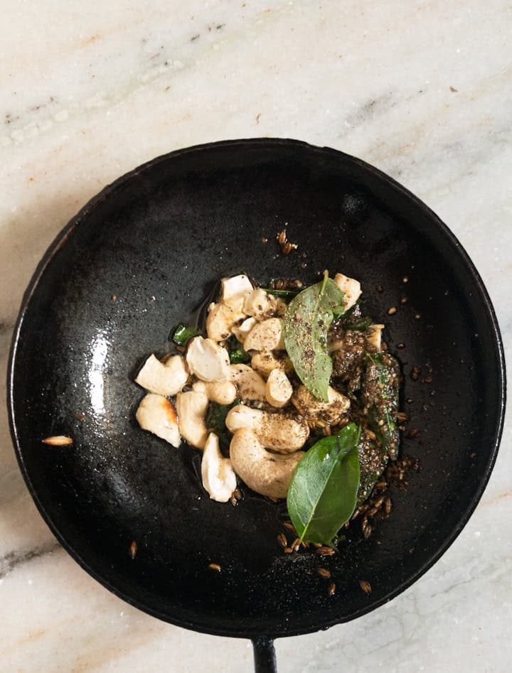 Cashew tadka with curry leaves, cumin, black pepper, and spices sizzling in a black pan.