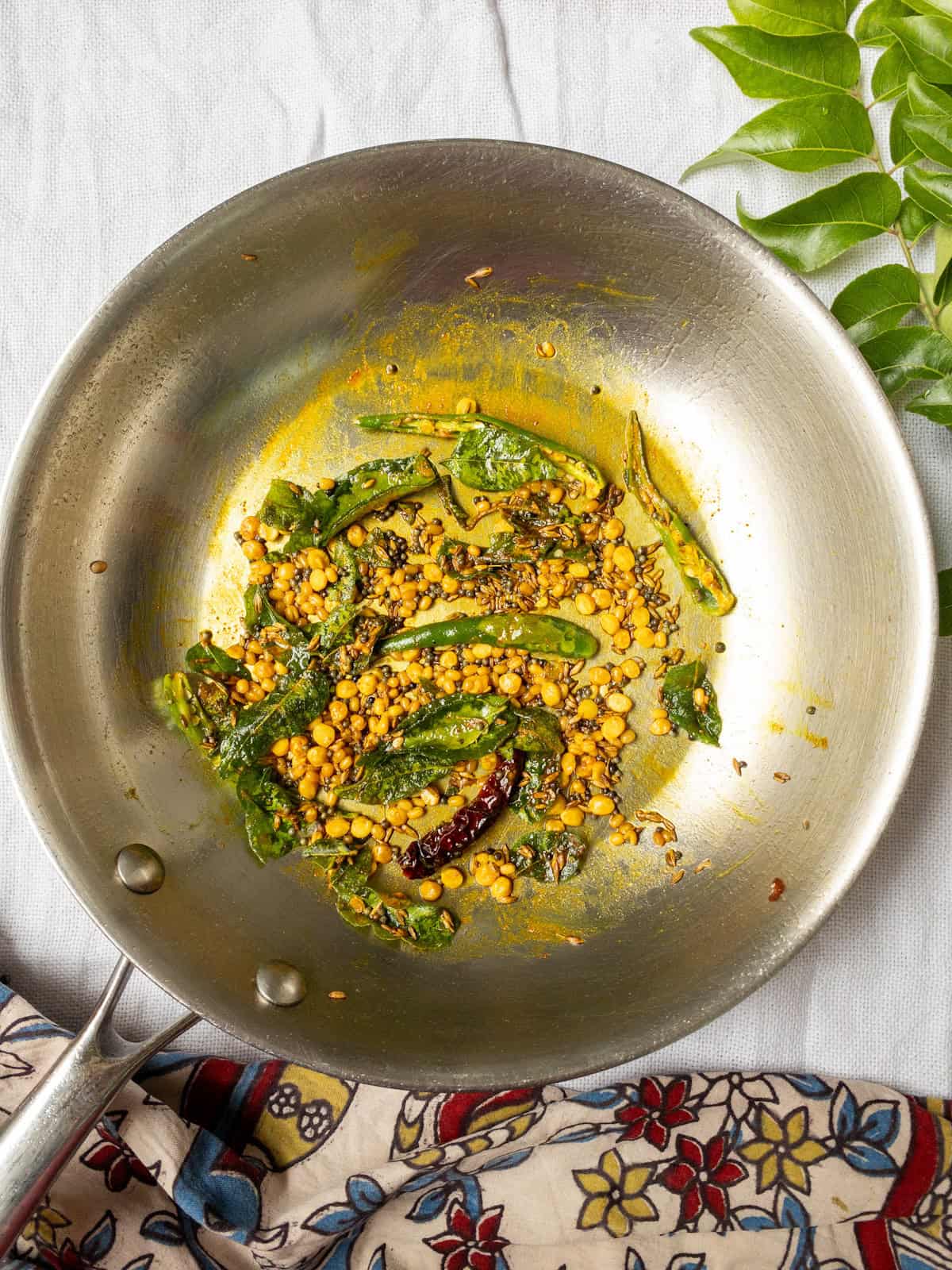 South Indian tadka with curry leaves, green chilies, red chili, mustard seeds, cumin seeds, and chana dal frying in a pan with turmeric.