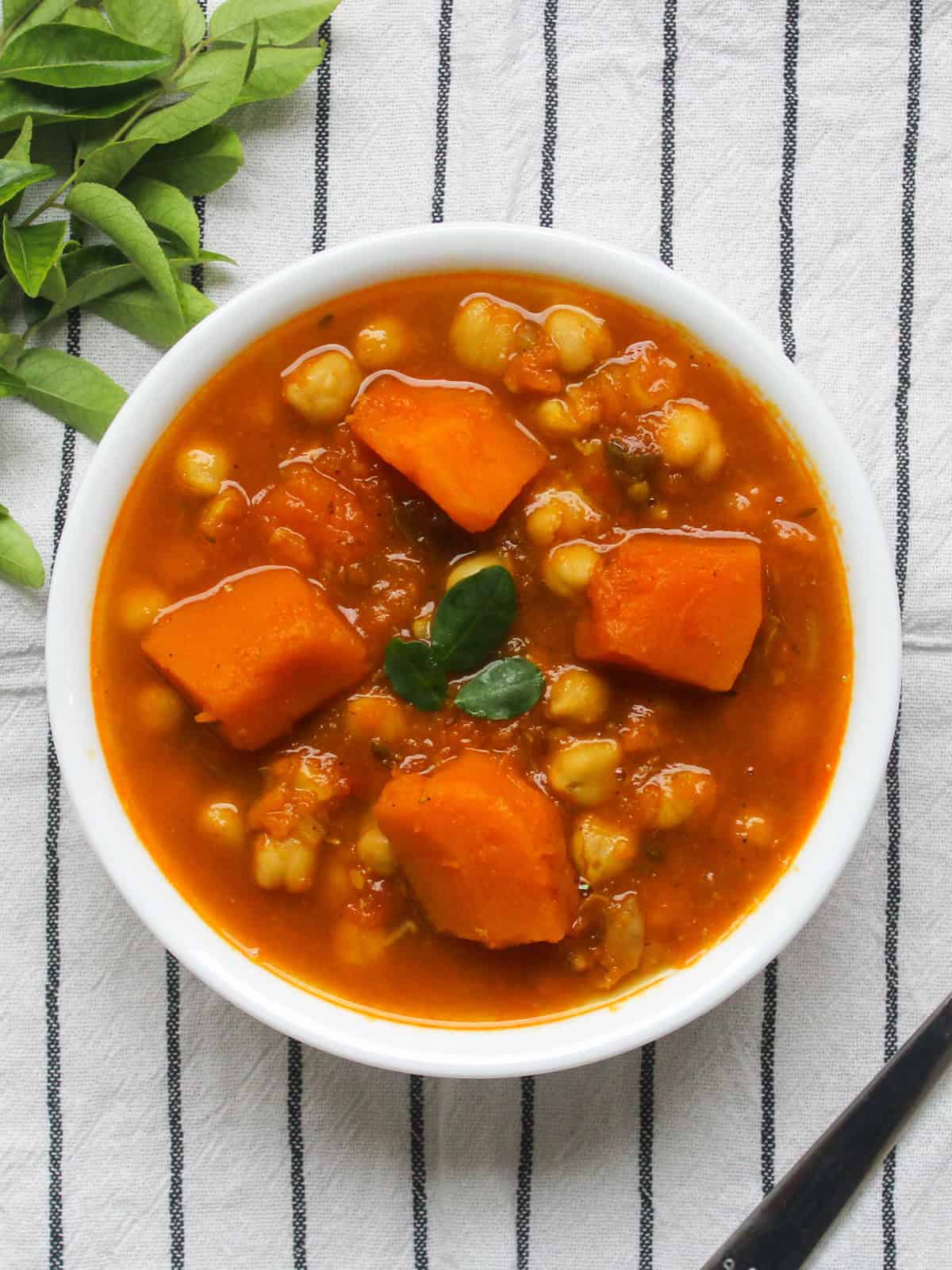 Bowl of cozy pumpkin chickpea curry served on striped napkin, garnished with fresh curry leaves.