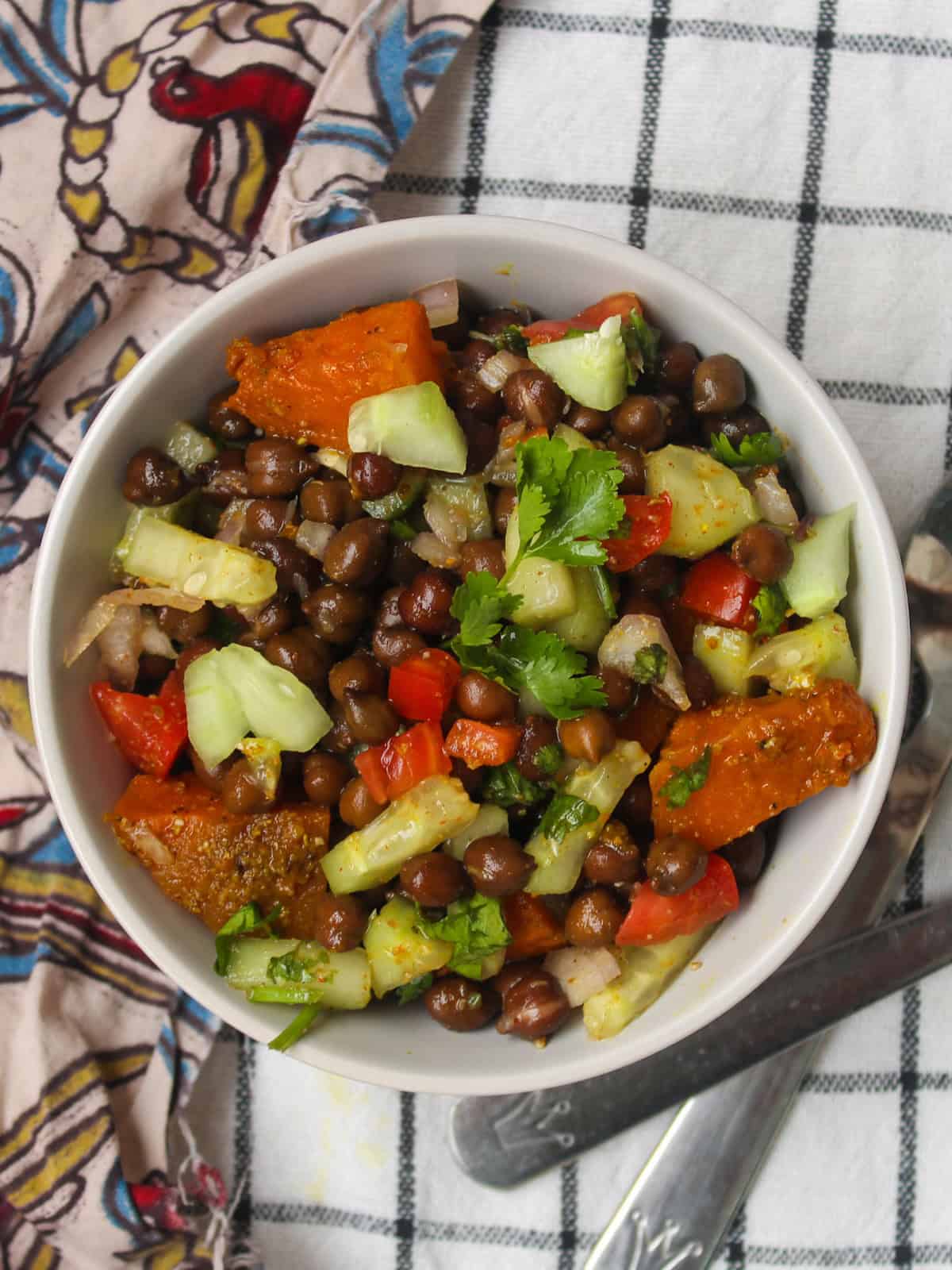 Bowl of roasted pumpkin and black chickpea salad with cucumber, tomato, onion, and cilantro served on a patterned cloth background.
