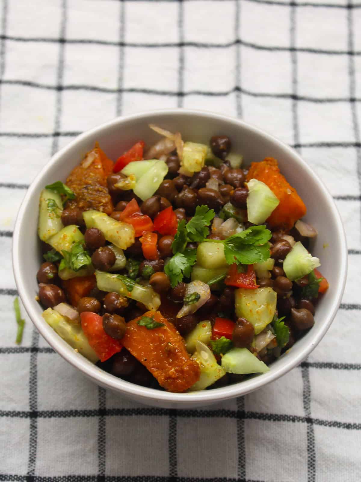 Indian roasted pumpkin and black chickpea salad with colorful vegetables in a white bowl placed on a checkered cloth.