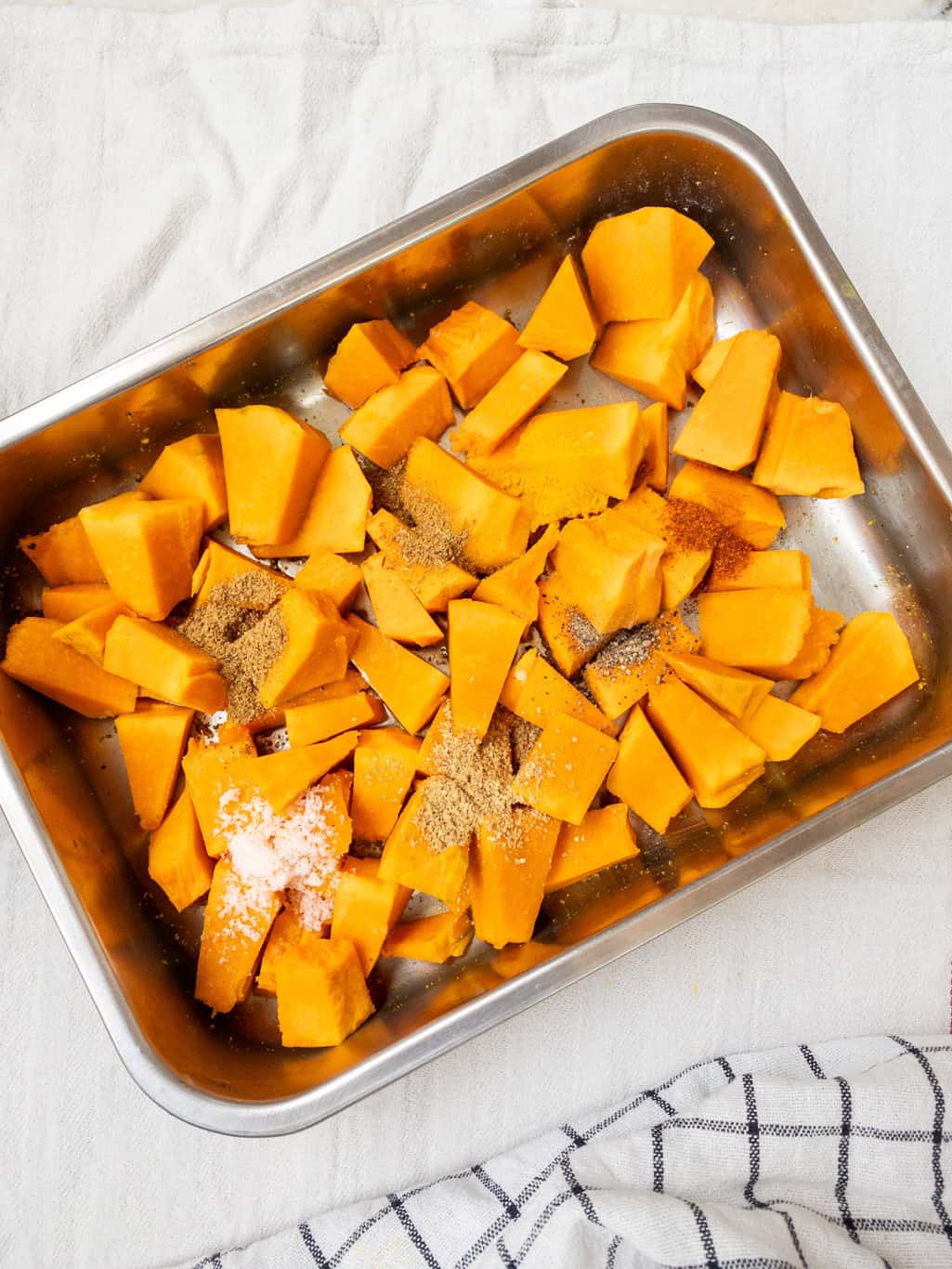 Cubes of pumpkin in a baking tray with Indian spices like salt, cumin, and coriander sprinkled on top before mixing for roasting.