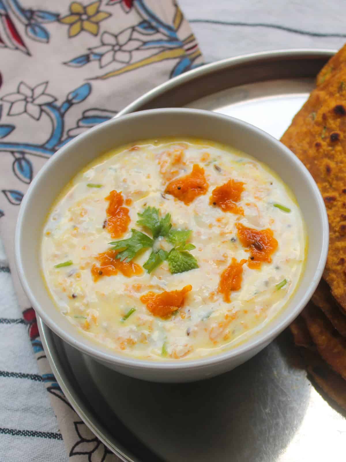 Creamy pumpkin raita served in a white bowl, garnished with cooked pumpkin pieces and cilantro, with pumpkin paratha on the side.