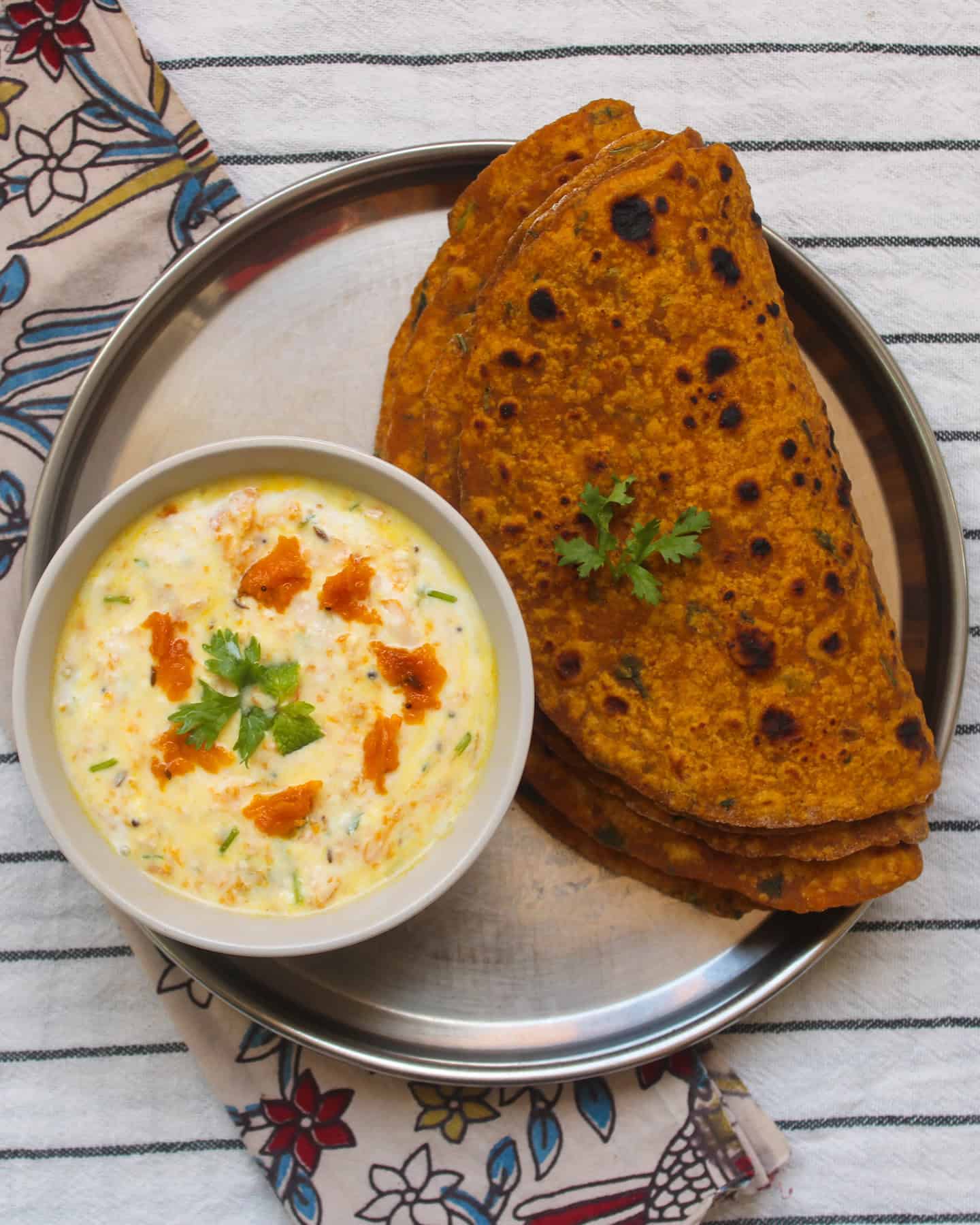 Pumpkin raita served in a bowl alongside stacked pumpkin parathas on a stainless steel plate, placed on a striped cloth.