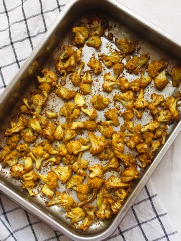 A tray of Indian spiced roasted cauliflower placed on a checkered kitchen towel, showing evenly browned, crisp florets.