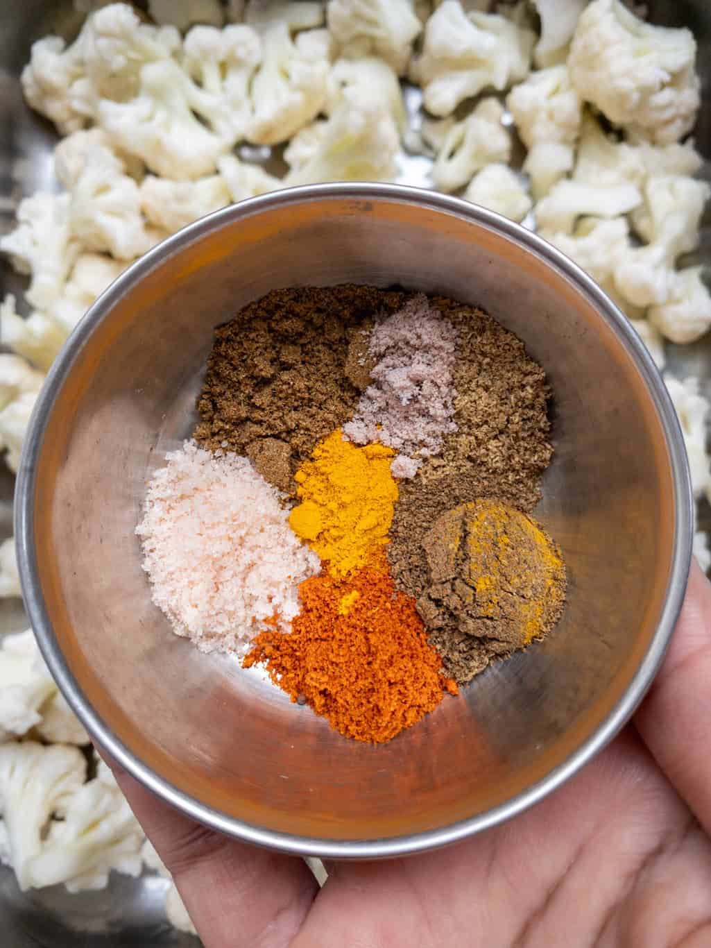 A small bowl filled with individual Indian spices-turmeric, red chilli powder, cumin, coriander, garam masala, and salt-held over a tray of cauliflower.
