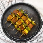 Top view of tandoori tofu tikka skewers placed on a black cast-iron pan, with a white checkered cloth underneath.
