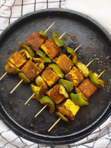 Top view of tandoori tofu tikka skewers placed on a black cast-iron pan, with a white checkered cloth underneath.