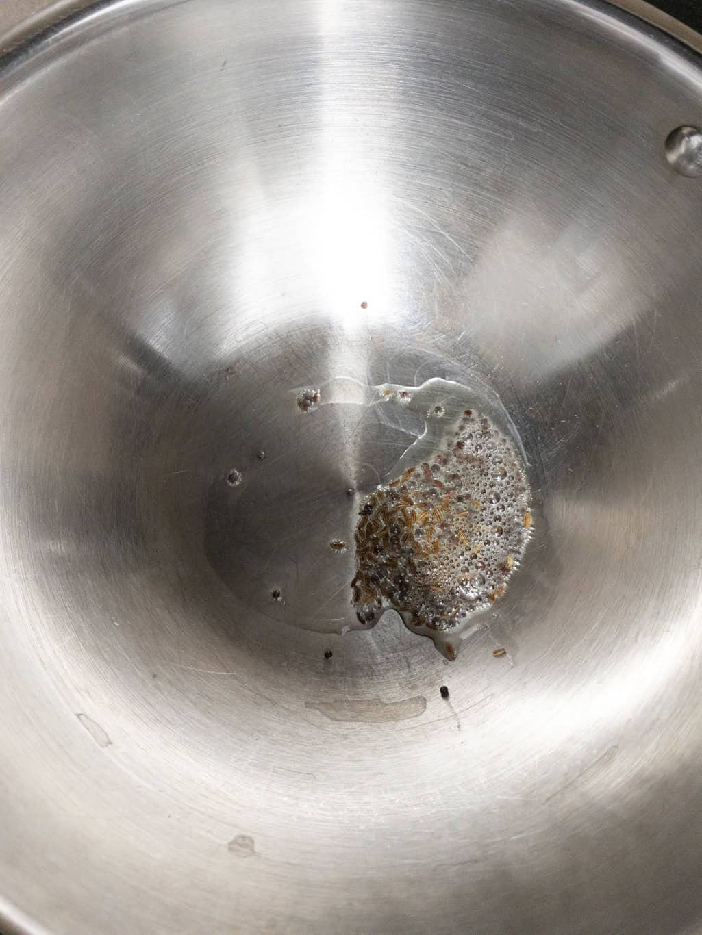 Mustard seeds and cumin seeds sizzling in oil in a steel pan.