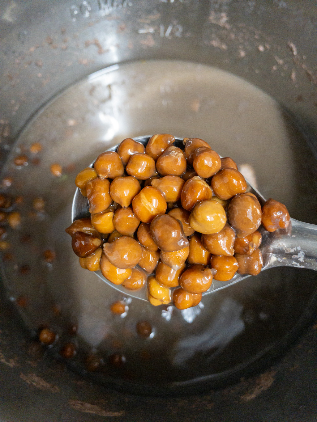 Cooked brown chickpeas held on a spoon above the cooking liquid.