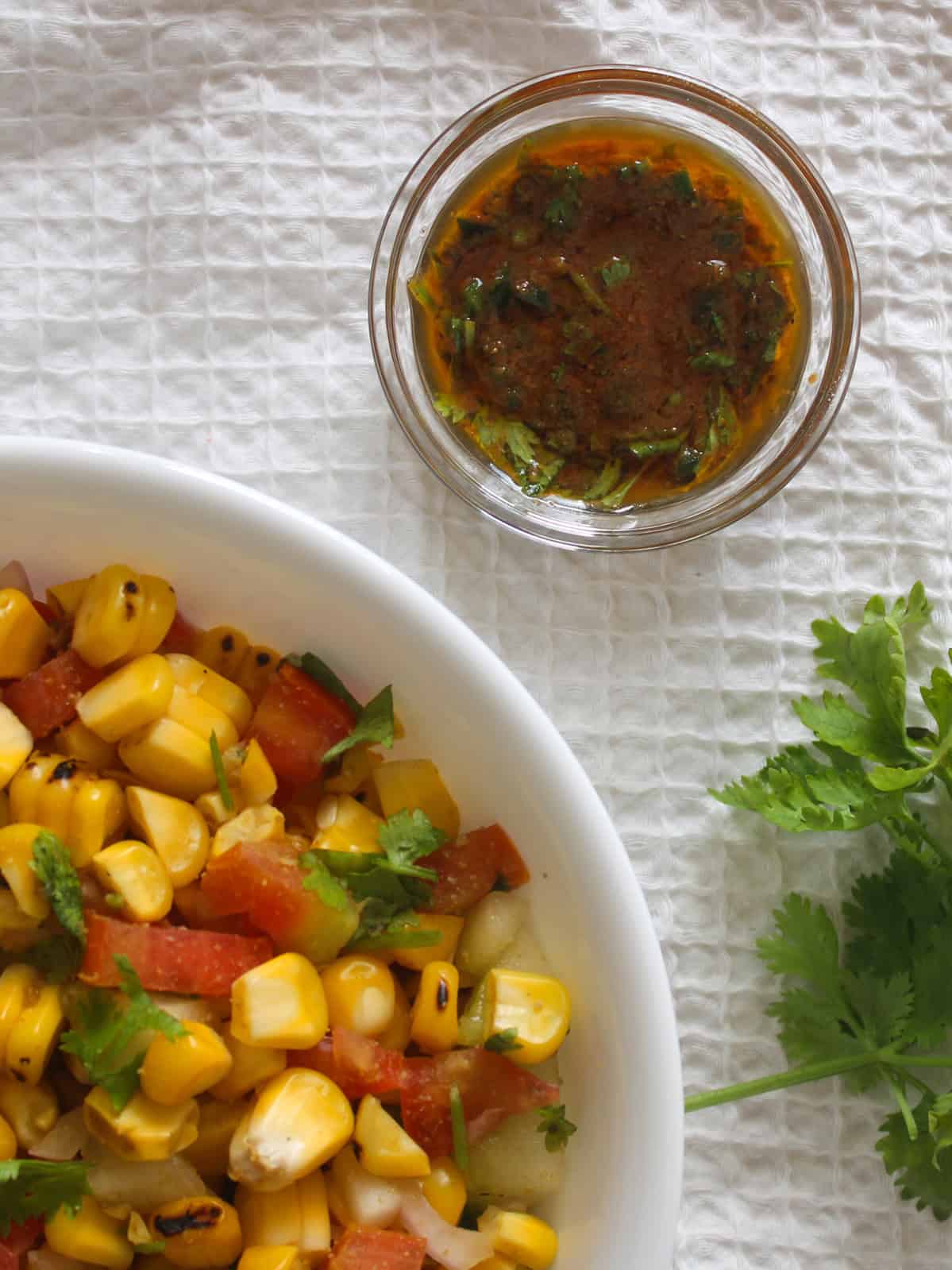 Chaat masala lemon dressing in a small glass bowl beside corn salad with tomato, cucumber, onion, and coriander.