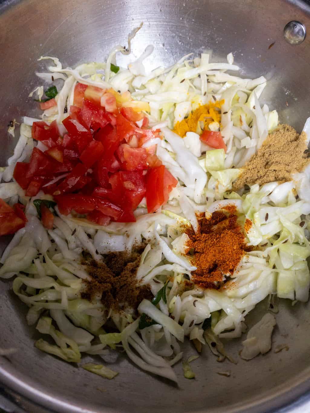 Shredded cabbage with chopped tomato and spice powders added in a steel pan.