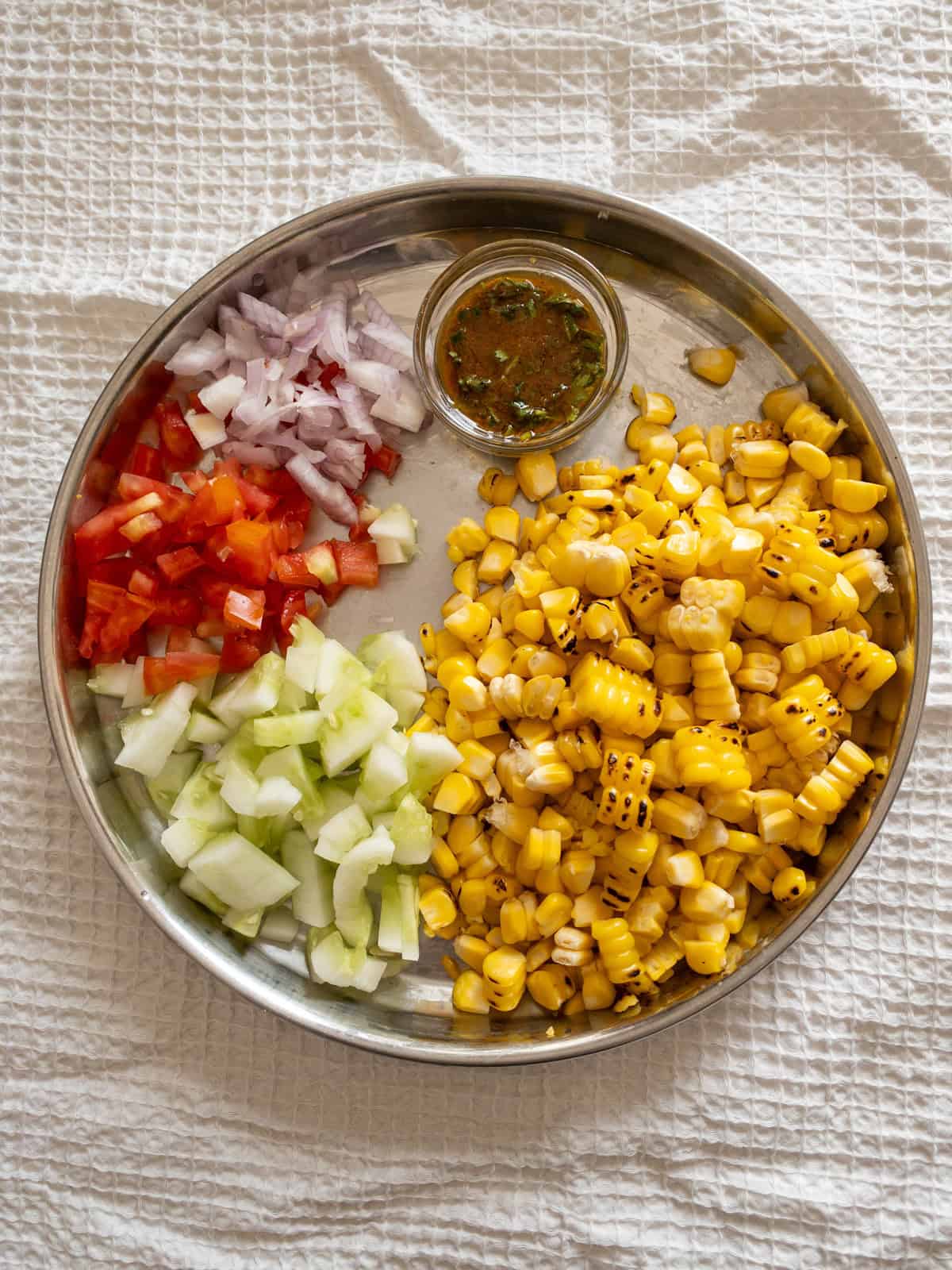 Grilled corn, chopped cucumber, tomato, onion, and prepared dressing arranged on a steel plate.