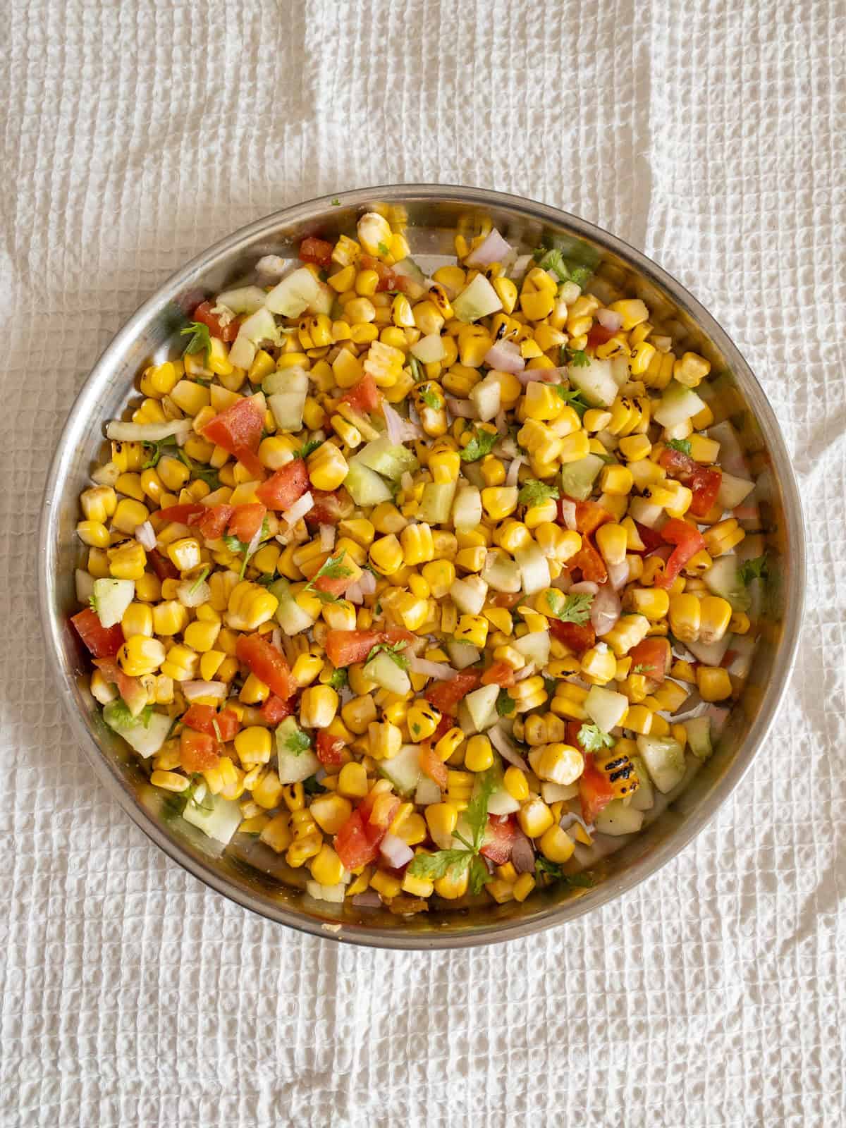 Grilled corn salad tossed with cucumber, tomato, onion, coriander, and chaat spices in a steel bowl.