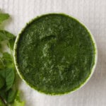Thick Indian green sauce in a white bowl with fresh cilantro and mint beside it.