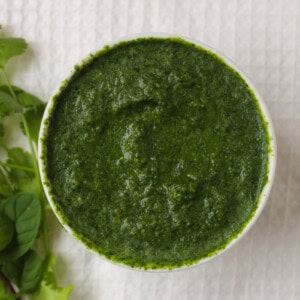 Thick Indian green sauce in a white bowl with fresh cilantro and mint beside it.