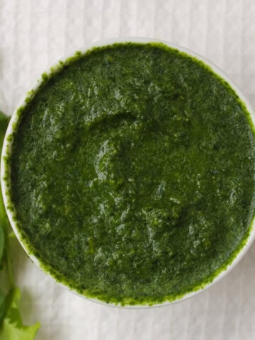 Thick Indian green sauce in a white bowl with fresh cilantro and mint beside it.