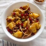 White bowl of Indian potato salad showing tender potato pieces, sliced onion, pomegranate seeds, and crushed peanuts.
