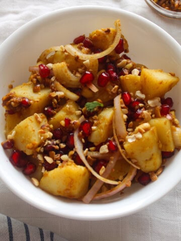 White bowl of Indian potato salad showing tender potato pieces, sliced onion, pomegranate seeds, and crushed peanuts.