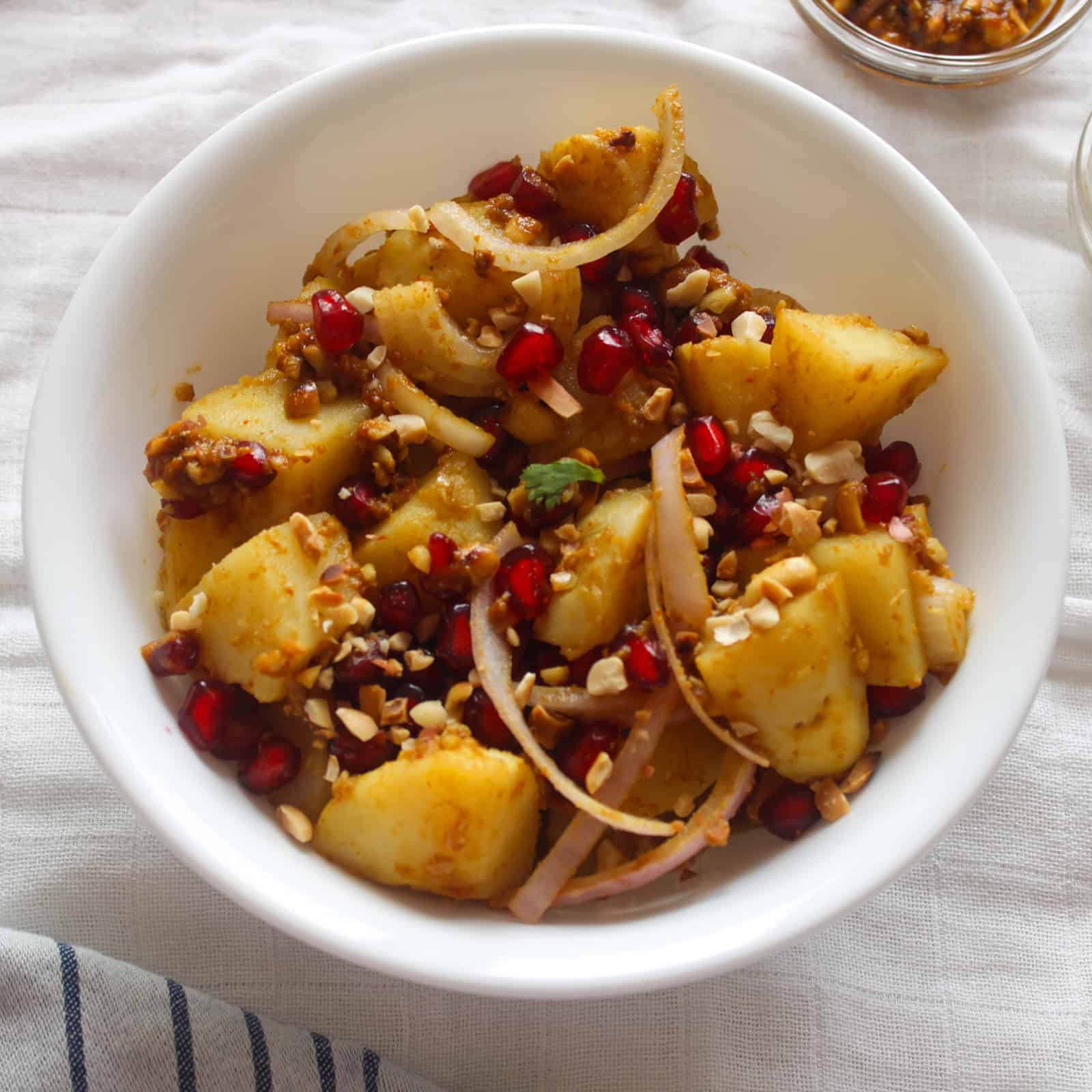 White bowl of Indian potato salad showing tender potato pieces, sliced onion, pomegranate seeds, and crushed peanuts.
