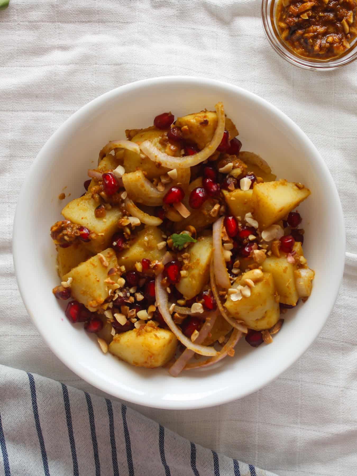 Overhead serving bowl filled with Indian potato salad, pomegranate seeds, sliced onion, crushed peanuts, and tamarind dressing.